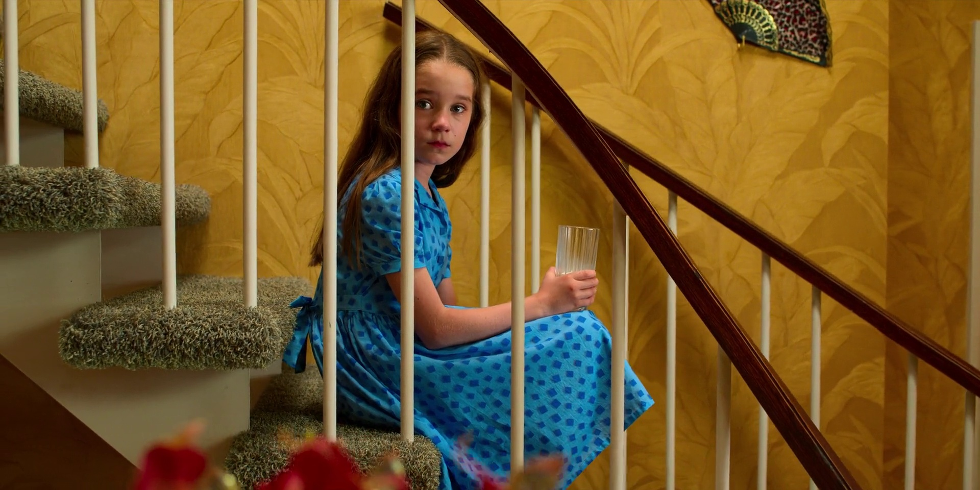 Fig 7: A young girl with long brown hair sits on the carpeted stairs, wearing a blue dress with a pattern of small dots. She holds a clear glass in her right hand and looks directly at the camera with a neutral expression. The stairs have white railings and the carpet is a light green color. The background features a yellow wall with a leafy pattern and a decorative fan hanging on the wall. The lighting is soft and natural, coming from the left side of the image, casting gentle shadows. The style of the image is a photograph. There are no other characters or text visible in the image..